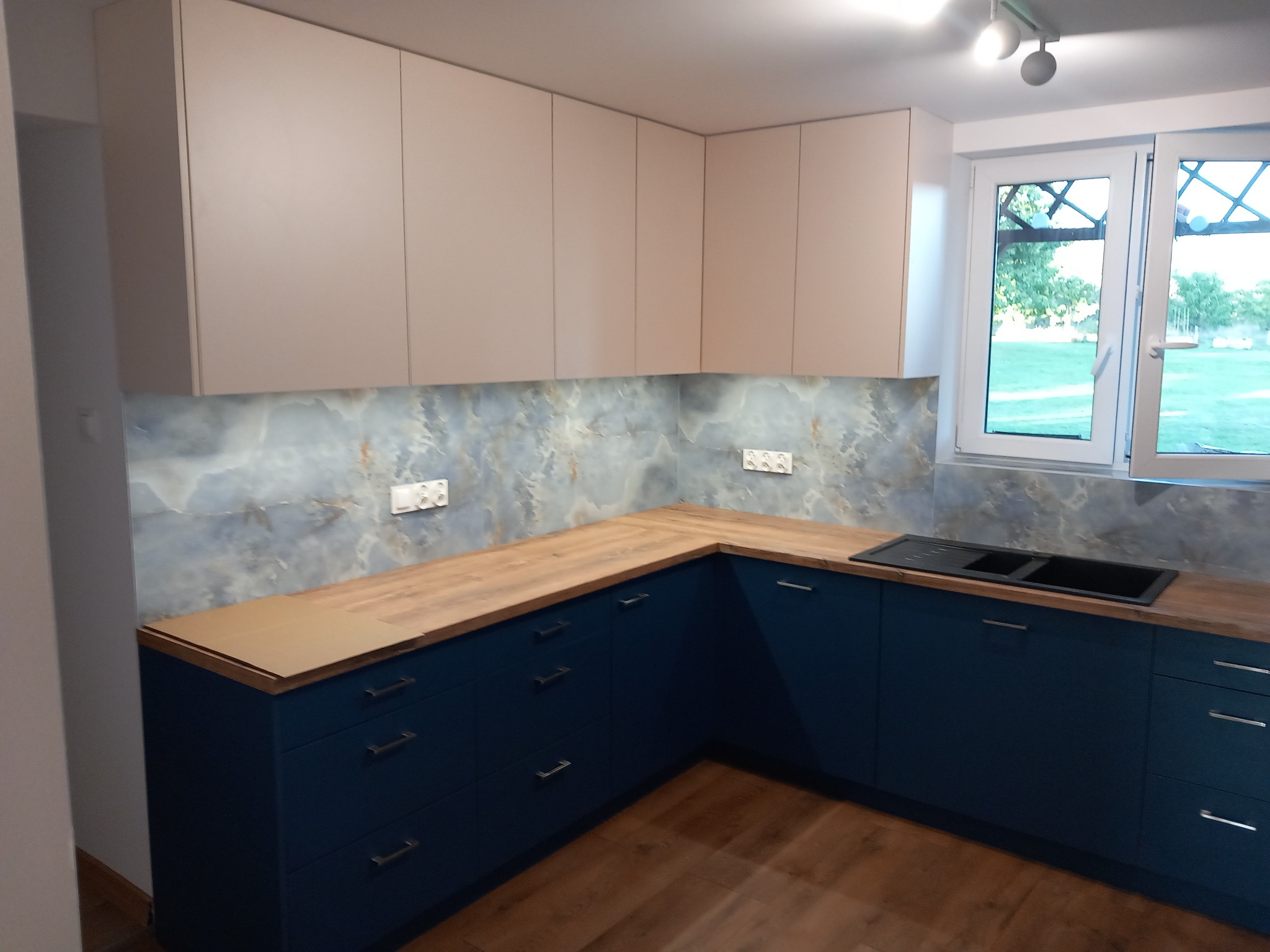 Nowoczesna kuchnia z granatowymi szafkami dolnymi, drewnianymi blatami i beżowymi szafkami górnymi, marmurowy backsplash, widok na ogród przez okno.