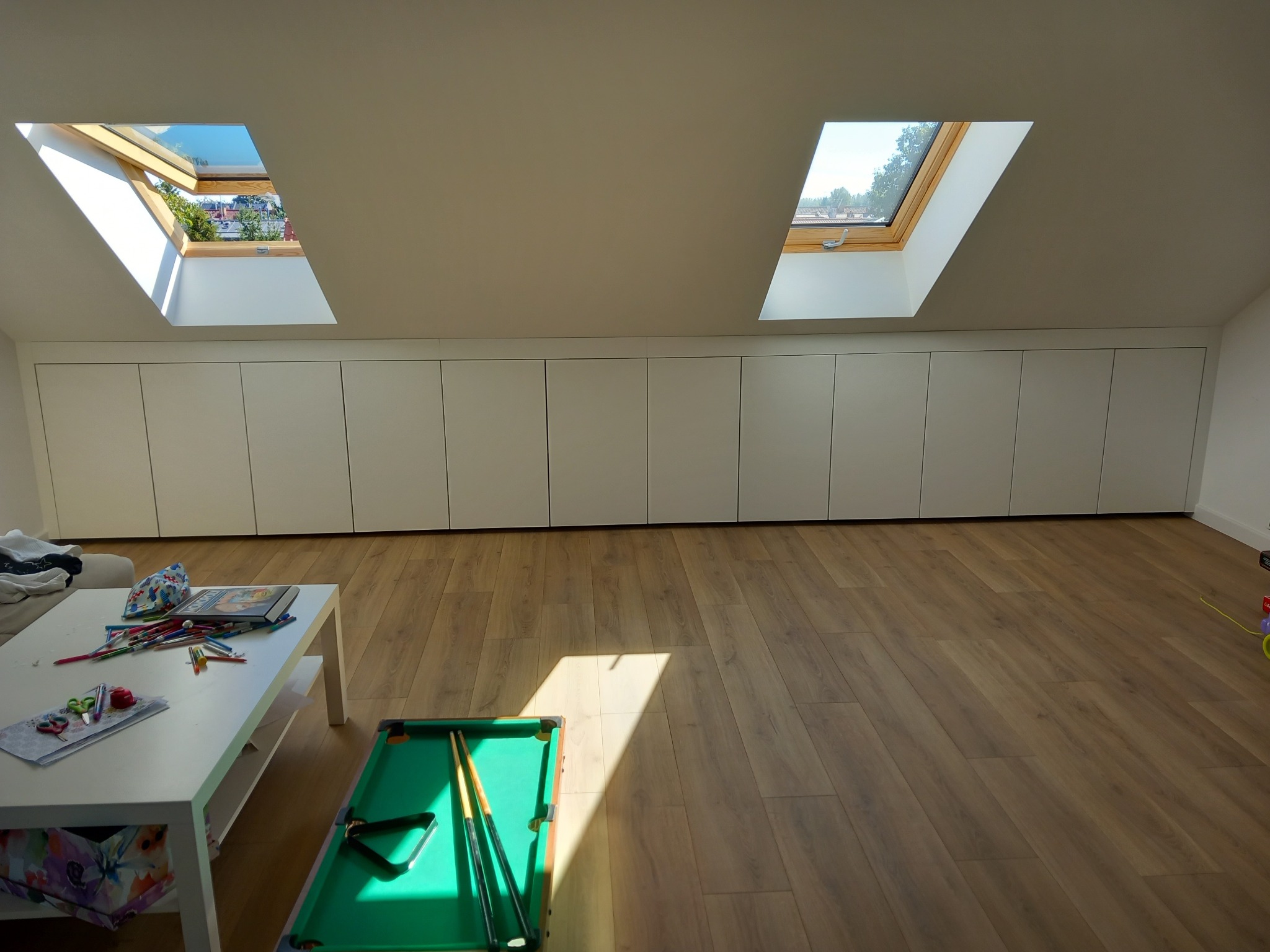 Białe szafy wnękowe pod skosami dachu z oknami dachowymi w pokoju z drewnianą podłogą, stolik z zabawkami i mały stół bilardowy.