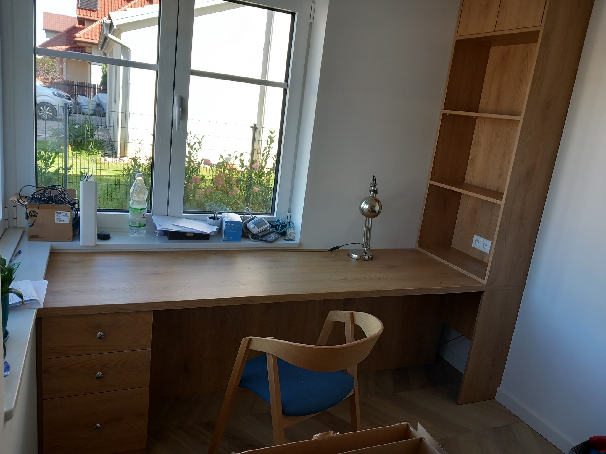 Drewniane biurko z szufladami i regałem, wbudowane pod oknem, z krzesłem z niebieskim siedziskiem, oświetlone srebrną lampką.