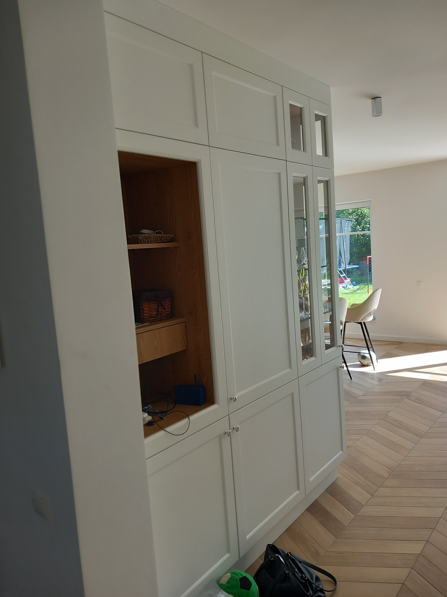 Biała zabudowa meblowa z przeszklonymi witrynami i otwartą półką z drewnianym wnętrzem, na podłodze parkiet w jodełkę, w tle widok na ogród przez okno.