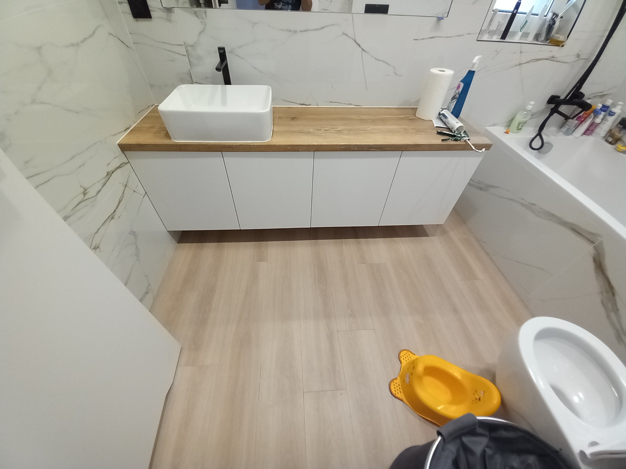 Nowoczesna łazienka z białą umywalką nablatową na drewnianym blacie, białe szafki pod umywalką, marmurowe płytki na ścianach, widoczny fragment wanny, toalety i nocnika dziecięcego na podłodze.