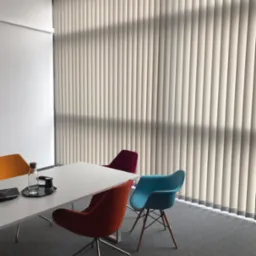 Beżowe, pionowe osłony okienne w biurze z nowoczesnym stołem konferencyjnym i kolorowymi krzesłami.