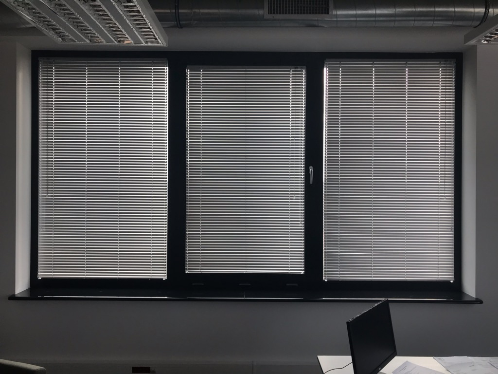 Trzyczęściowe okno z czarną ramą, osłonięte poziomymi listwami w kolorze srebrnym, widoczne fragmenty biurka z komputerem i dokumentami, oświetlenie sufitowe.