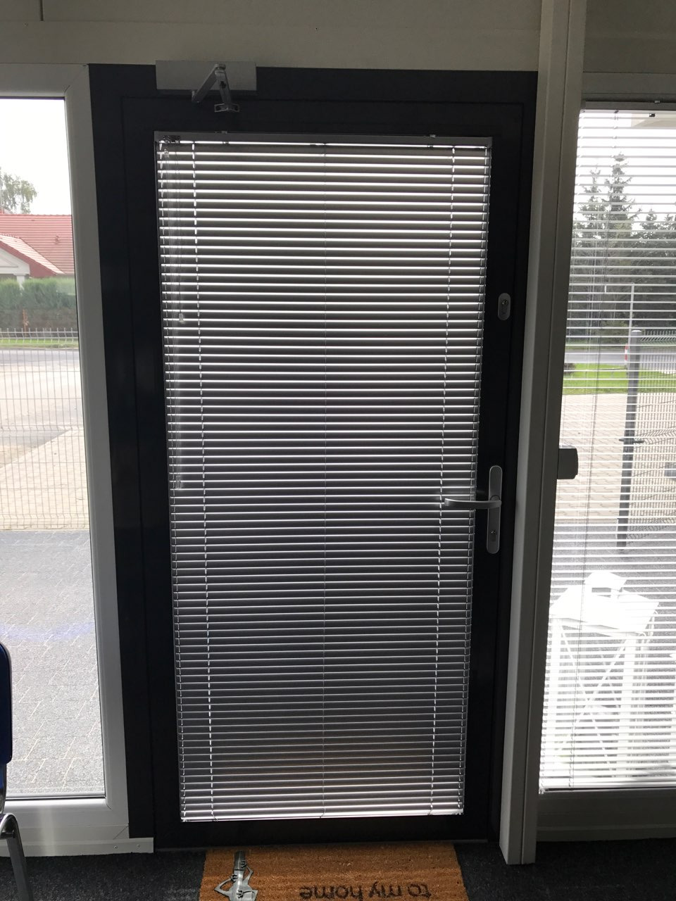 Zamknięte, poziome przesłony okienne w szarej ramie drzwiowej, widoczne przez przeszklone drzwi wejściowe z wycieraczką 'to my home'.