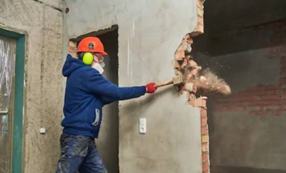 Pracownik w kasku i ochronnikach słuchu uderza młotem w ceglaną ścianę, wzbijając tumany pyłu, widoczne okablowanie i fragment sąsiedniego pomieszczenia.