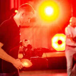 Mężczyzna obsługujący profesjonalną kamerę w intensywnym czerwonym oświetleniu, z parą w tle i dekoracyjnymi światłami.