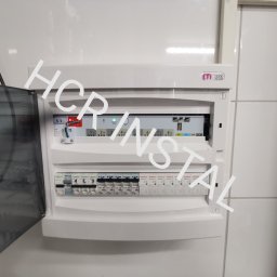 HCR Instal - Otwarta skrzynka elektryczna z zainstalowanymi bezpiecznikami i modułem sterującym, zamontowana na białej ścianie z płytek.