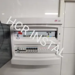 Otwarta skrzynka elektryczna z zainstalowanymi bezpiecznikami i modułem sterującym, zamontowana na białej ścianie z płytek.