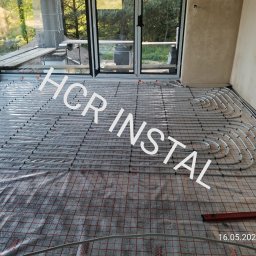 HCR Instal - Rozłożona instalacja ogrzewania podłogowego na folii z siatką w nowym budynku z dużym oknem balkonowym, widoczne rury grzewcze i poziomica na podłodze.