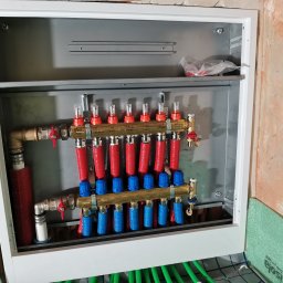 HCR Instal - Rozdzielacz ogrzewania podłogowego w metalowej szafce, widoczne rury w izolacji czerwonej i niebieskiej, zielone rury ogrzewania podłogowego na podłodze z izolacją. Ściany w stanie surowym.