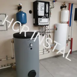 Kompleksowa instalacja grzewcza w kotłowni: piec, zasobniki CWU, naczynia przeponowe, rozdzielacze, orurowanie z armaturą, skrzynka elektryczna na ścianie, szare płytki podłogowe.