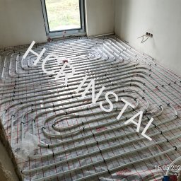 HCR Instal - Białe rury instalacji ogrzewania podłogowego ułożone na folii izolacyjnej z czerwonym nadrukiem kratki, widoczne fragmenty ścian w surowym stanie i okno.