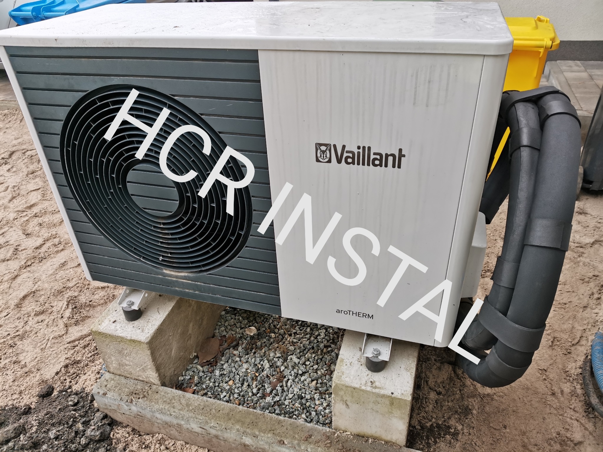 Zewnętrzna jednostka pompy ciepła Vaillant aroTHERM, zamontowana na betonowych blokach, widoczne izolowane rury i fragment żółtego pojemnika na śmieci w tle.