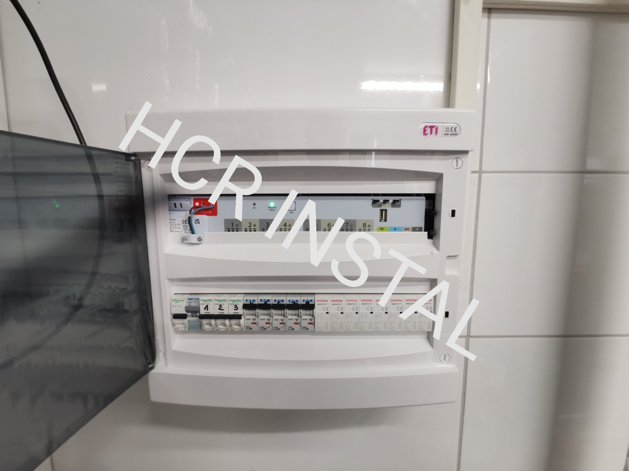 Otwarta skrzynka elektryczna z zainstalowanymi bezpiecznikami i modułem sterującym, zamontowana na białej ścianie z płytek.