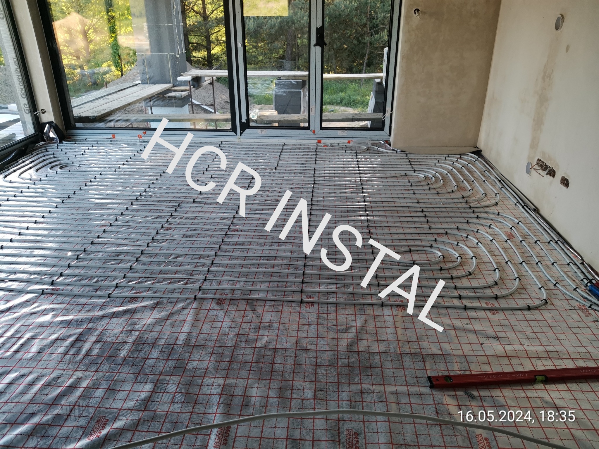 Rozłożona instalacja ogrzewania podłogowego na folii z siatką w nowym budynku z dużym oknem balkonowym, widoczne rury grzewcze i poziomica na podłodze.