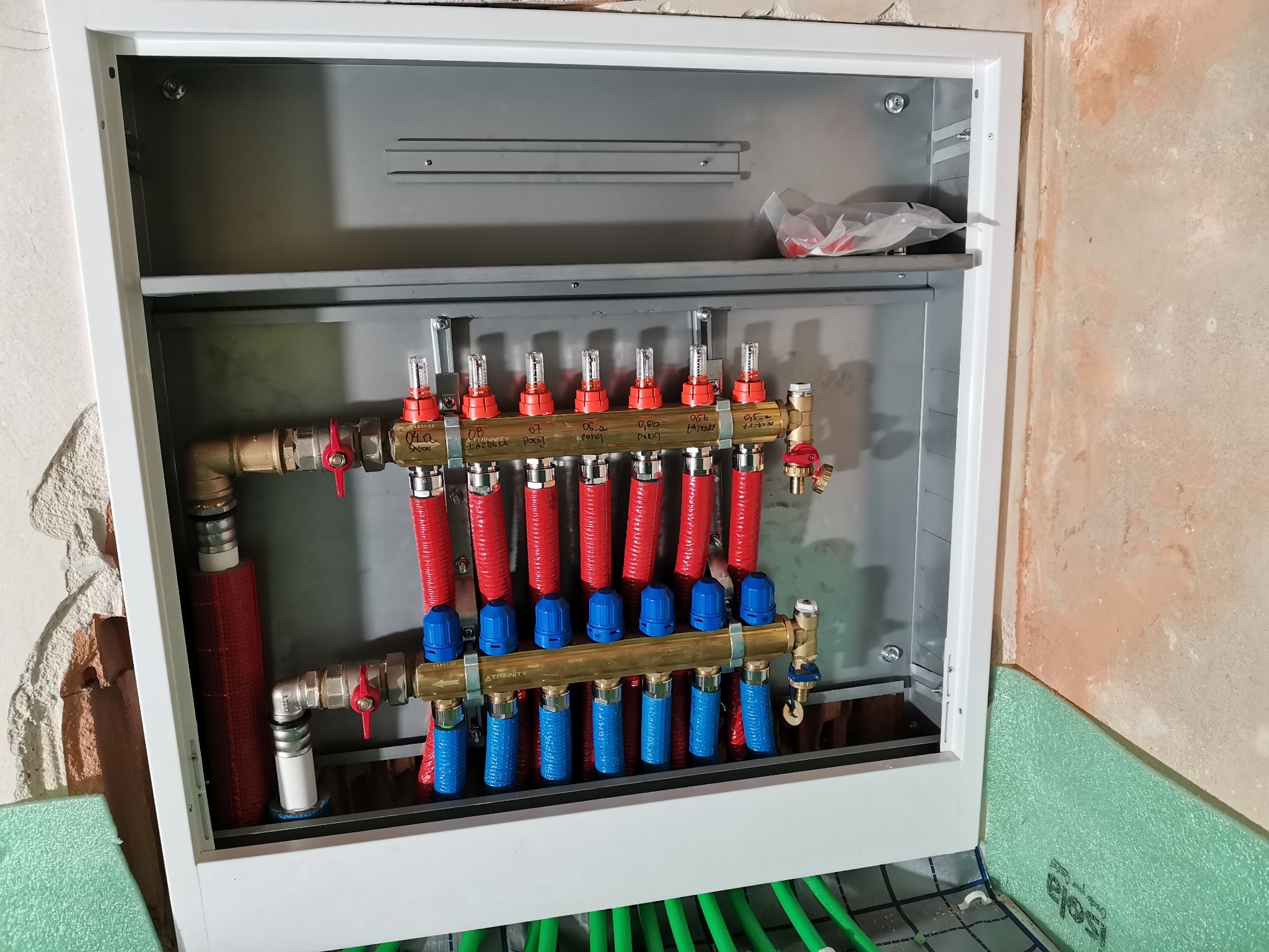 Rozdzielacz ogrzewania podłogowego w metalowej szafce, widoczne rury w izolacji czerwonej i niebieskiej, zielone rury ogrzewania podłogowego na podłodze z izolacją. Ściany w stanie surowym.