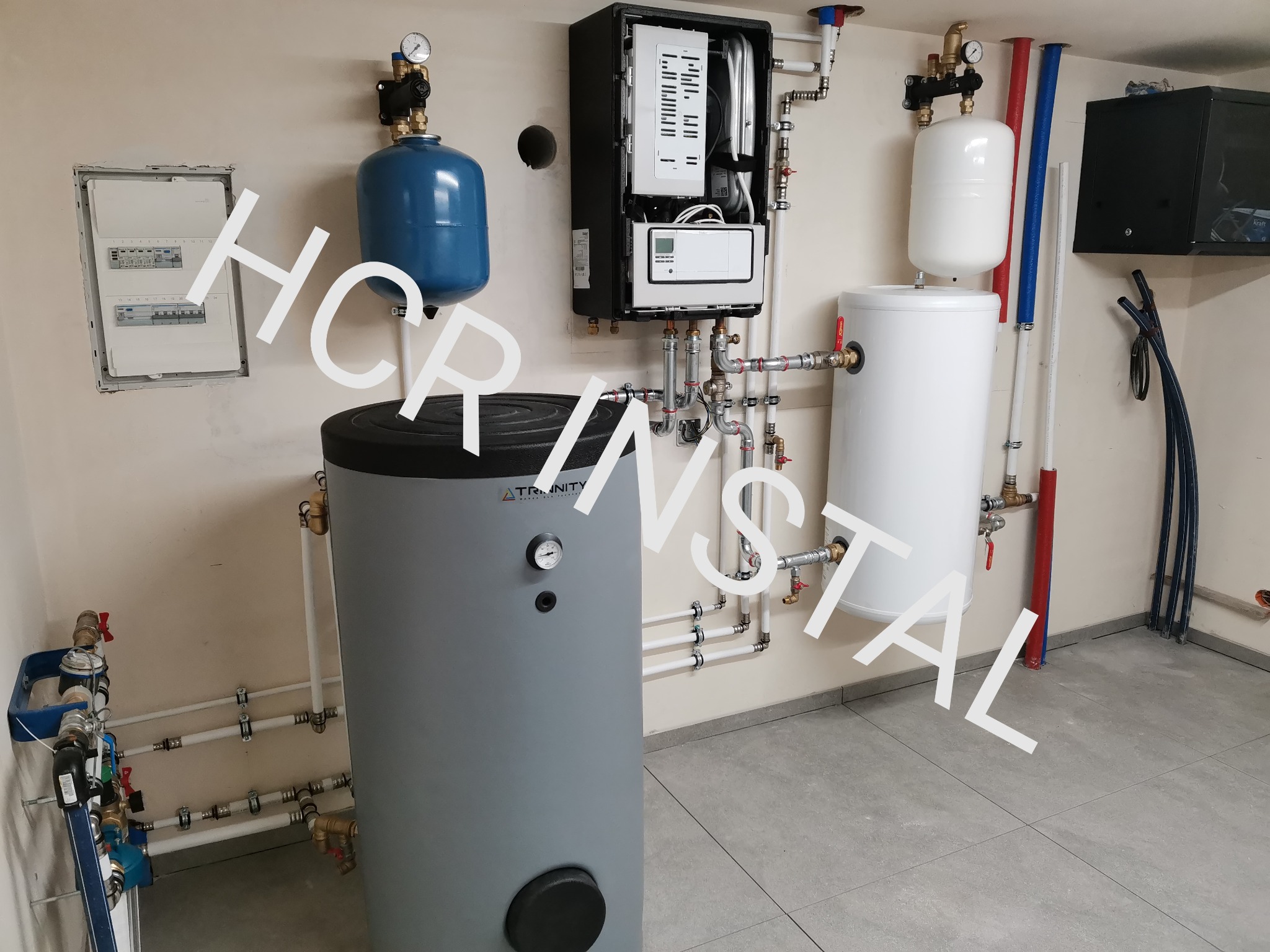 Kompleksowa instalacja grzewcza w kotłowni: piec, zasobniki CWU, naczynia przeponowe, rozdzielacze, orurowanie z armaturą, skrzynka elektryczna na ścianie, szare płytki podłogowe.