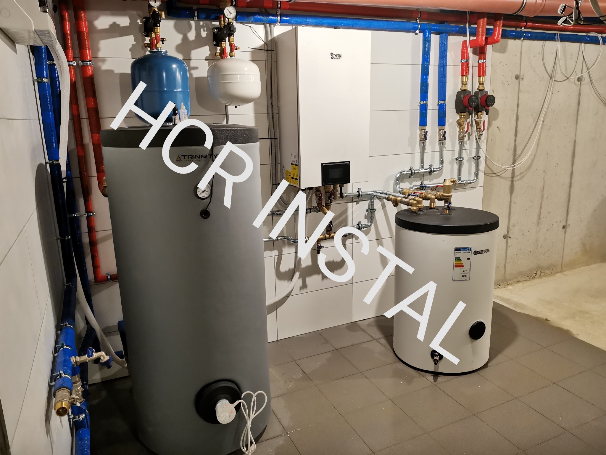 Nowoczesna kotłownia z białym piecem gazowym, szarym zasobnikiem ciepłej wody użytkowej marki Trinity i białym zasobnikiem Kospel, widoczne liczne rury w kolorach niebieskim i czerwonym oraz armatura.