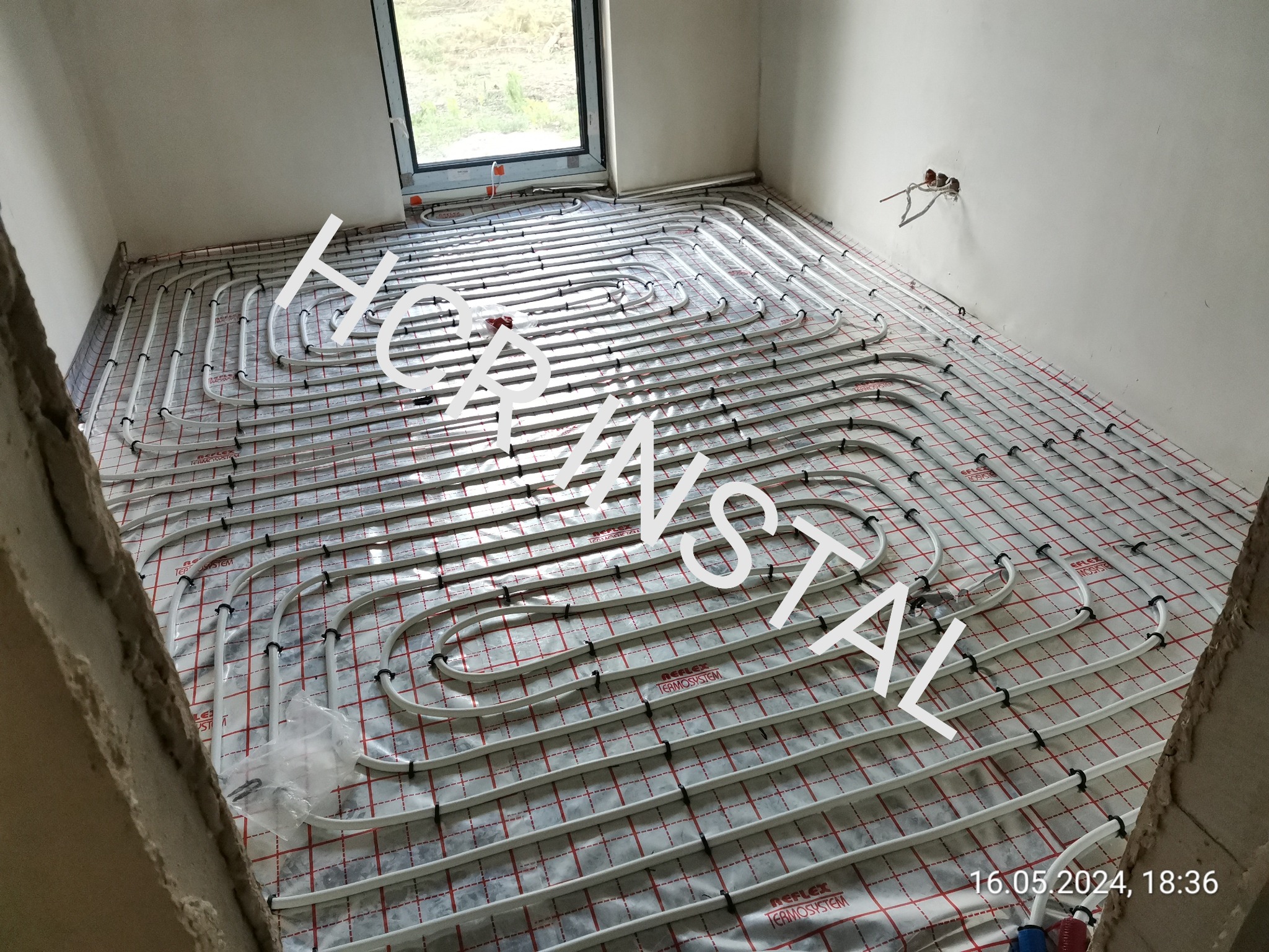 Białe rury instalacji ogrzewania podłogowego ułożone na folii izolacyjnej z czerwonym nadrukiem kratki, widoczne fragmenty ścian w surowym stanie i okno.