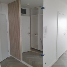 DARIUSZ LEWANDOWSKI - Nowoczesne wykończenie przedpokoju: lustro w zabudowie z drewnianymi lamelami, białe ściany i parkiet w jodełkę. Minimalistyczny design z widoczną szafą w tle.