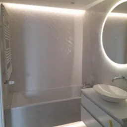 Nowoczesna łazienka z podświetlaną wanną, umywalką na szafce i okrągłym lustrem LED. Ściany wyłożone białymi płytkami w jodełkę, subtelne oświetlenie sufitowe.