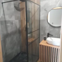 Nowoczesna łazienka: kabina walk-in z czarnymi profilami, umywalka nablatowa na drewnianej szafce z lamelami, okrągłe lustro podświetlane. Szare płytki imitujące kamień.