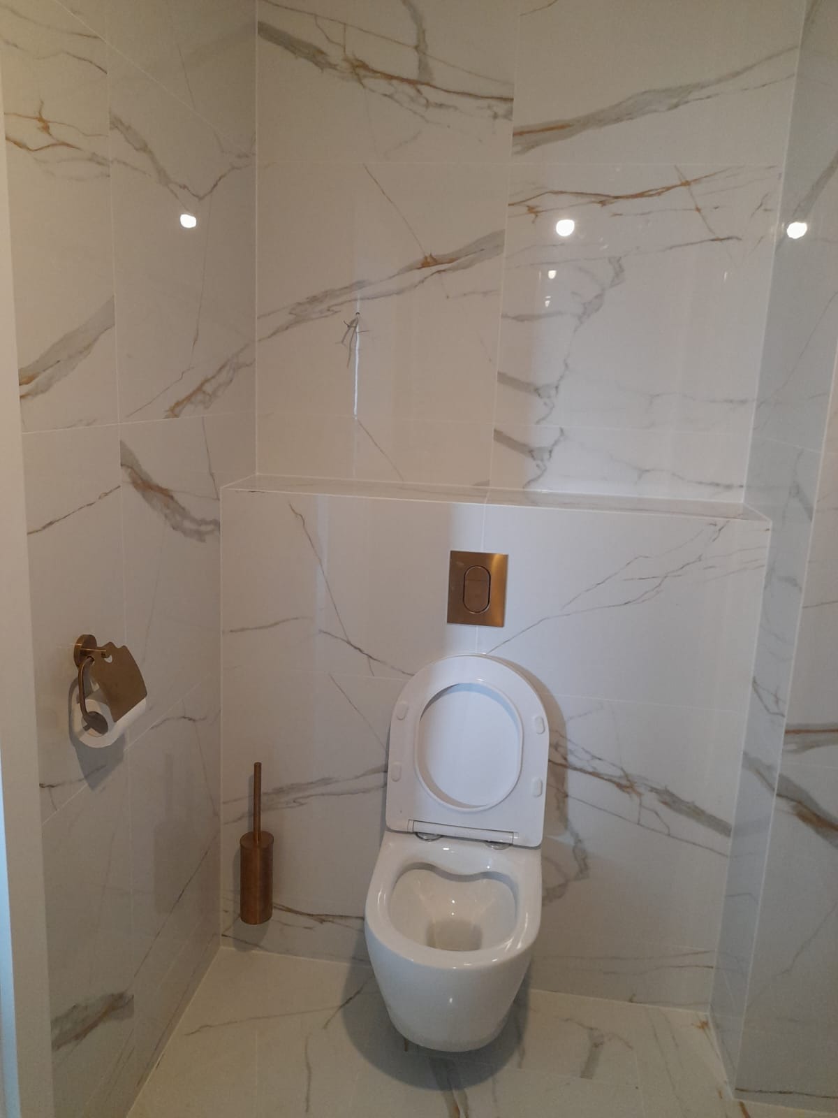 Wykończona łazienka z białymi płytkami imitującymi marmur z złotymi żyłami, wisząca toaleta i akcesoria w kolorze złotym.