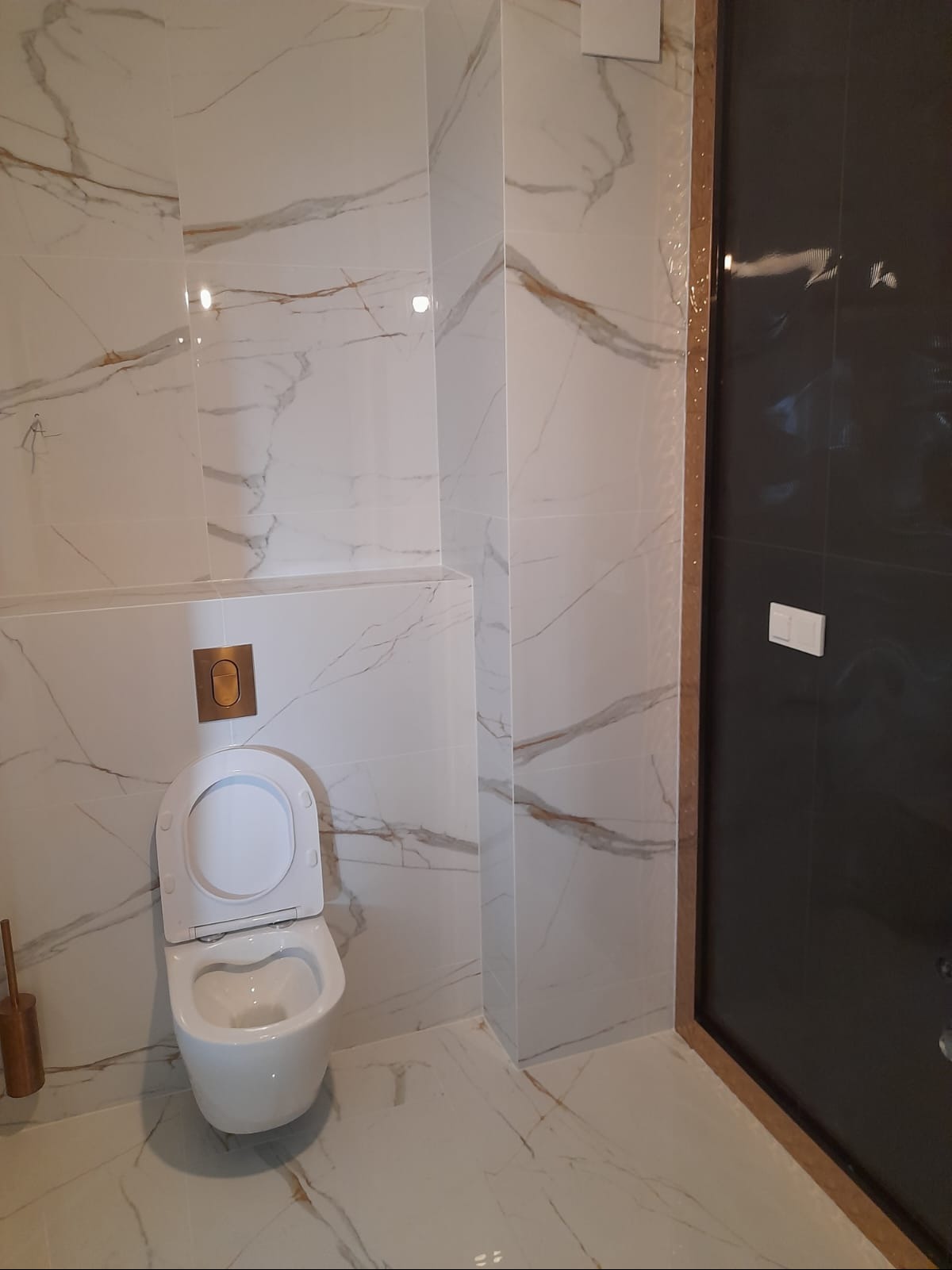 Nowoczesna łazienka z marmurowymi płytkami, wiszącą toaletą i czarnymi drzwiami kabiny prysznicowej. Złote akcenty na przycisku spłukiwania.