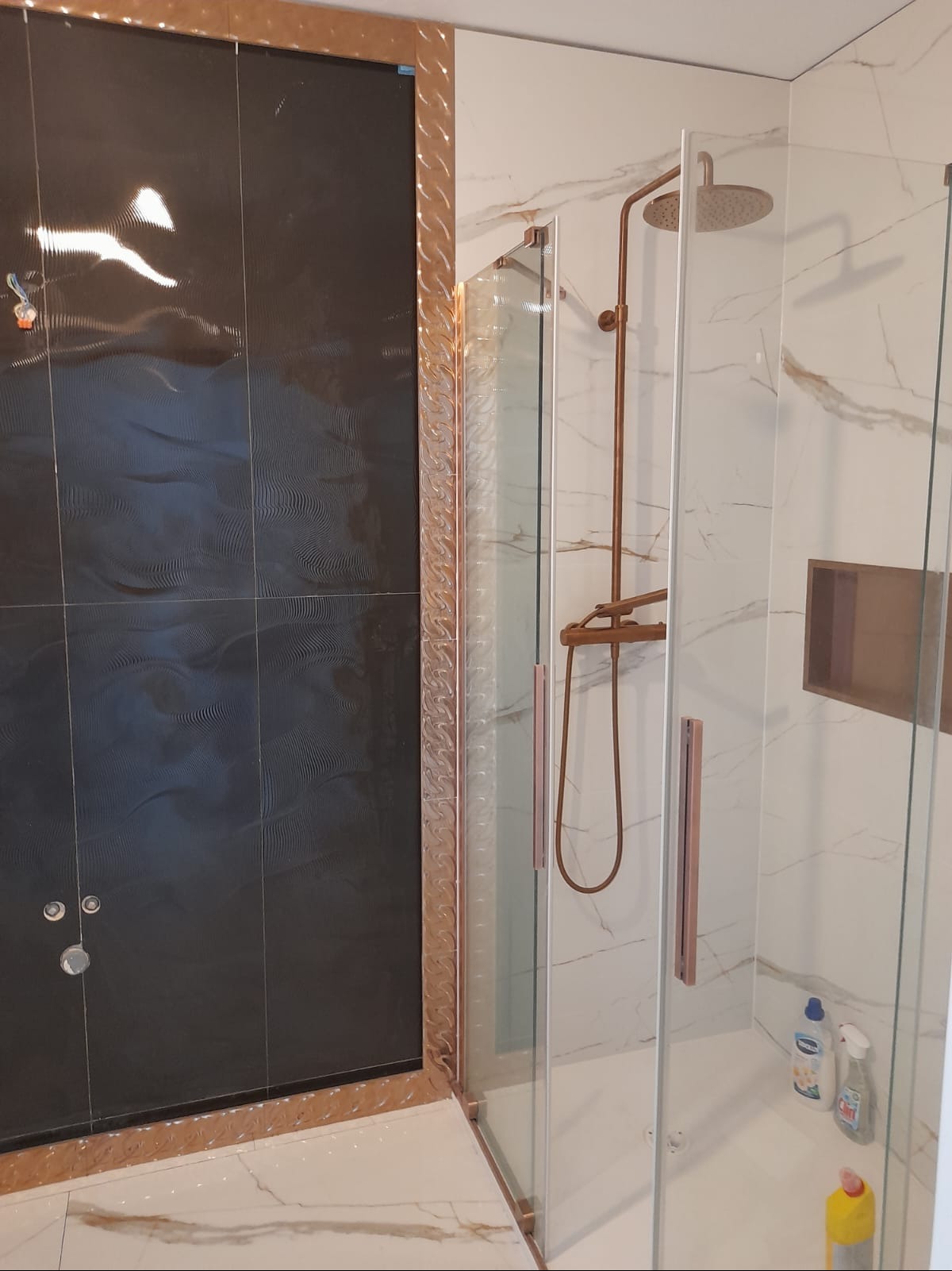 Nowoczesna kabina prysznicowa z czarnymi płytkami o falistej strukturze i złotymi elementami wykończeniowymi. Marmurowe płytki na ścianie z wnęką.