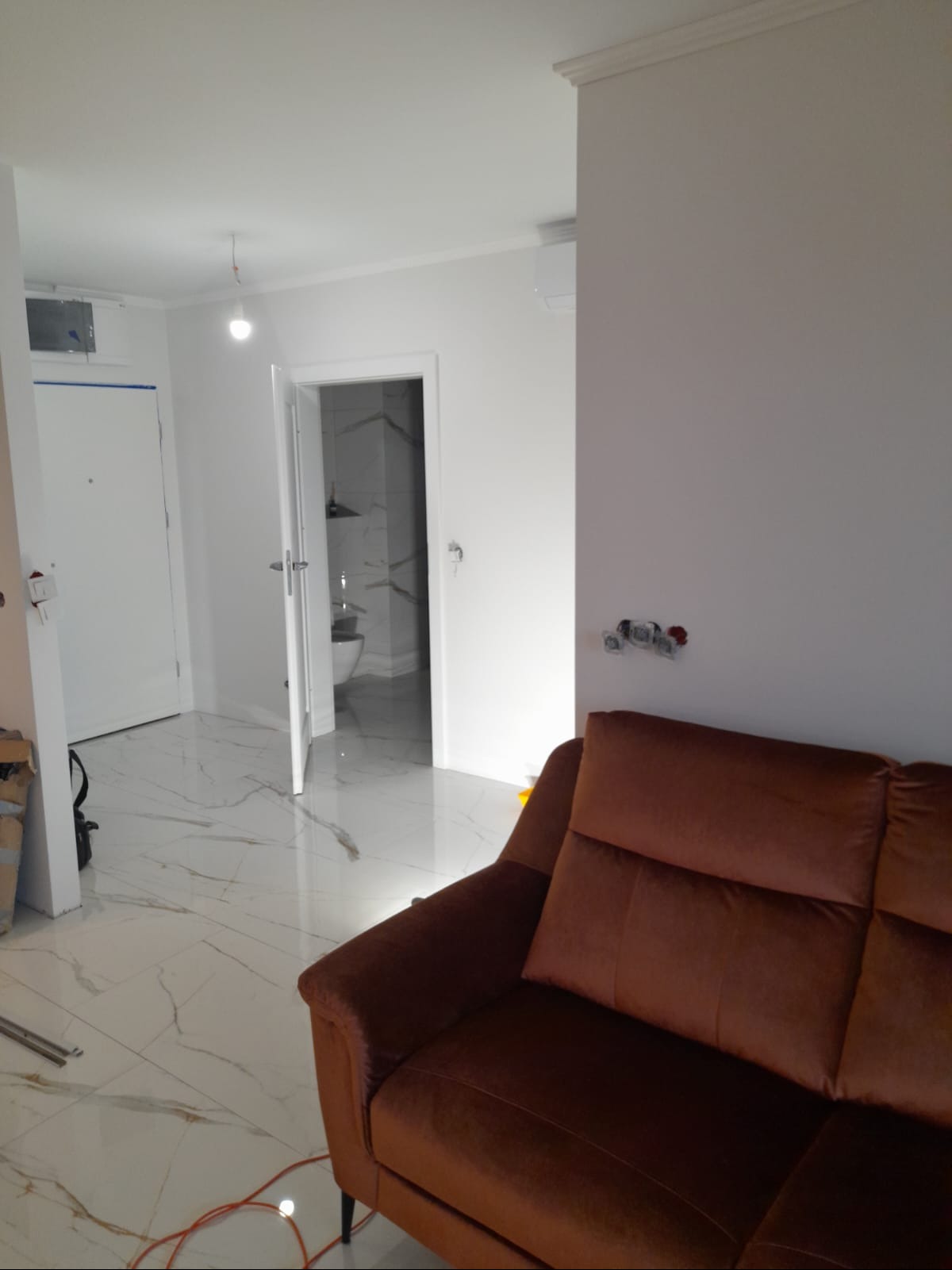 Jasne wnętrze po remoncie: białe ściany, marmurowe płytki podłogowe, widok na łazienkę z marmurowymi płytkami, brązowa sofa i wystające kable elektryczne.
