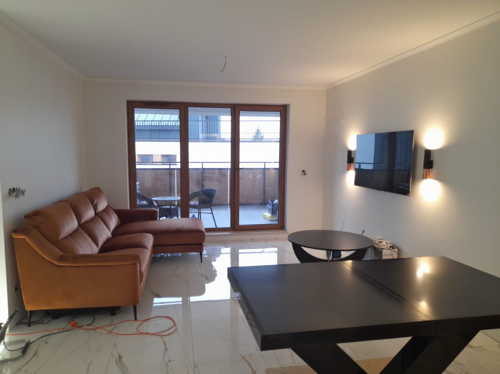 Jasne wnętrze salonu z marmurową podłogą, brązową sofą i wyjściem na balkon. Na ścianie zamontowany telewizor podświetlony ozdobnymi lampami.