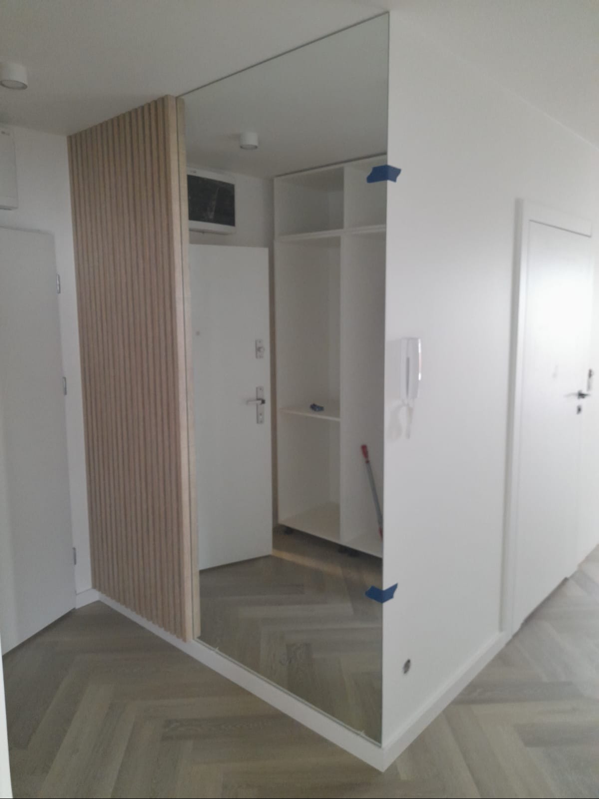 Nowoczesne wykończenie przedpokoju: lustro w zabudowie z drewnianymi lamelami, białe ściany i parkiet w jodełkę. Minimalistyczny design z widoczną szafą w tle.