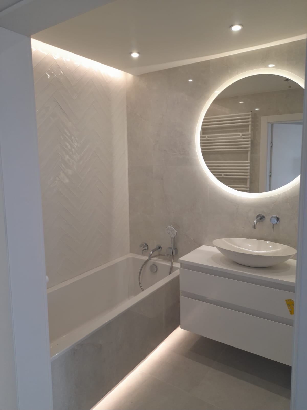 Elegancka łazienka w odcieniach szarości z białą armaturą i podświetlanym lustrem. Na ścianie płytki ułożone w jodełkę, dające ciekawy efekt wizualny.