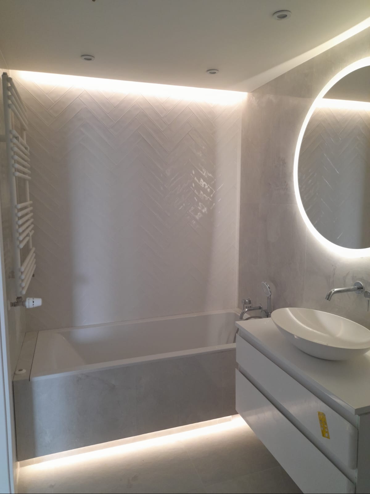 Nowoczesna łazienka z podświetlaną wanną, umywalką na szafce i okrągłym lustrem LED. Ściany wyłożone białymi płytkami w jodełkę, subtelne oświetlenie sufitowe.