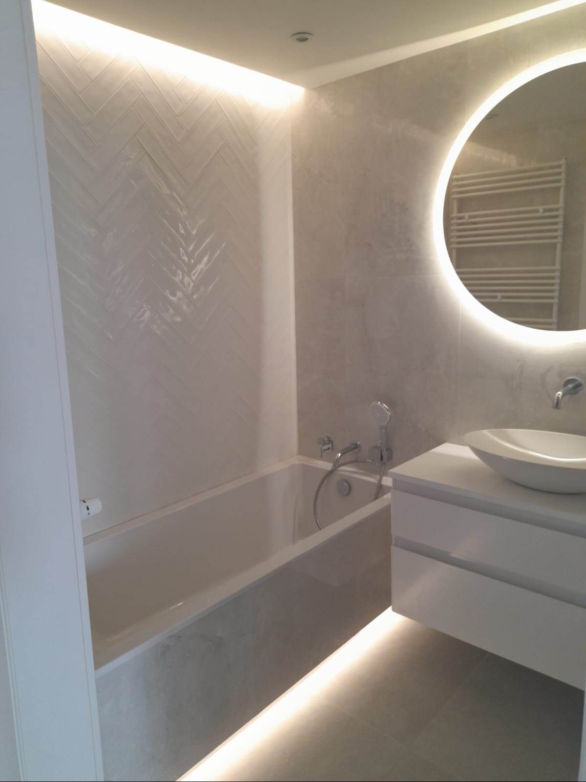 Nowoczesna łazienka z wanną, umywalką na szafce i okrągłym lustrem z podświetleniem LED. Ściana wyłożona białymi płytkami w jodełkę, oświetlenie pod wanną.