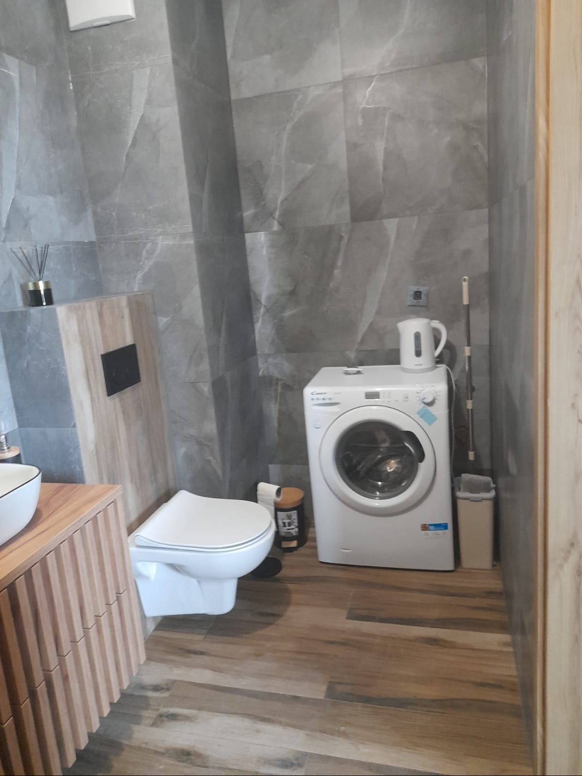 Nowoczesna łazienka z szarymi płytkami imitującymi marmur, drewnianą szafką pod umywalką i pralką. Na podłodze panele imitujące drewno. Minimalistyczny design.