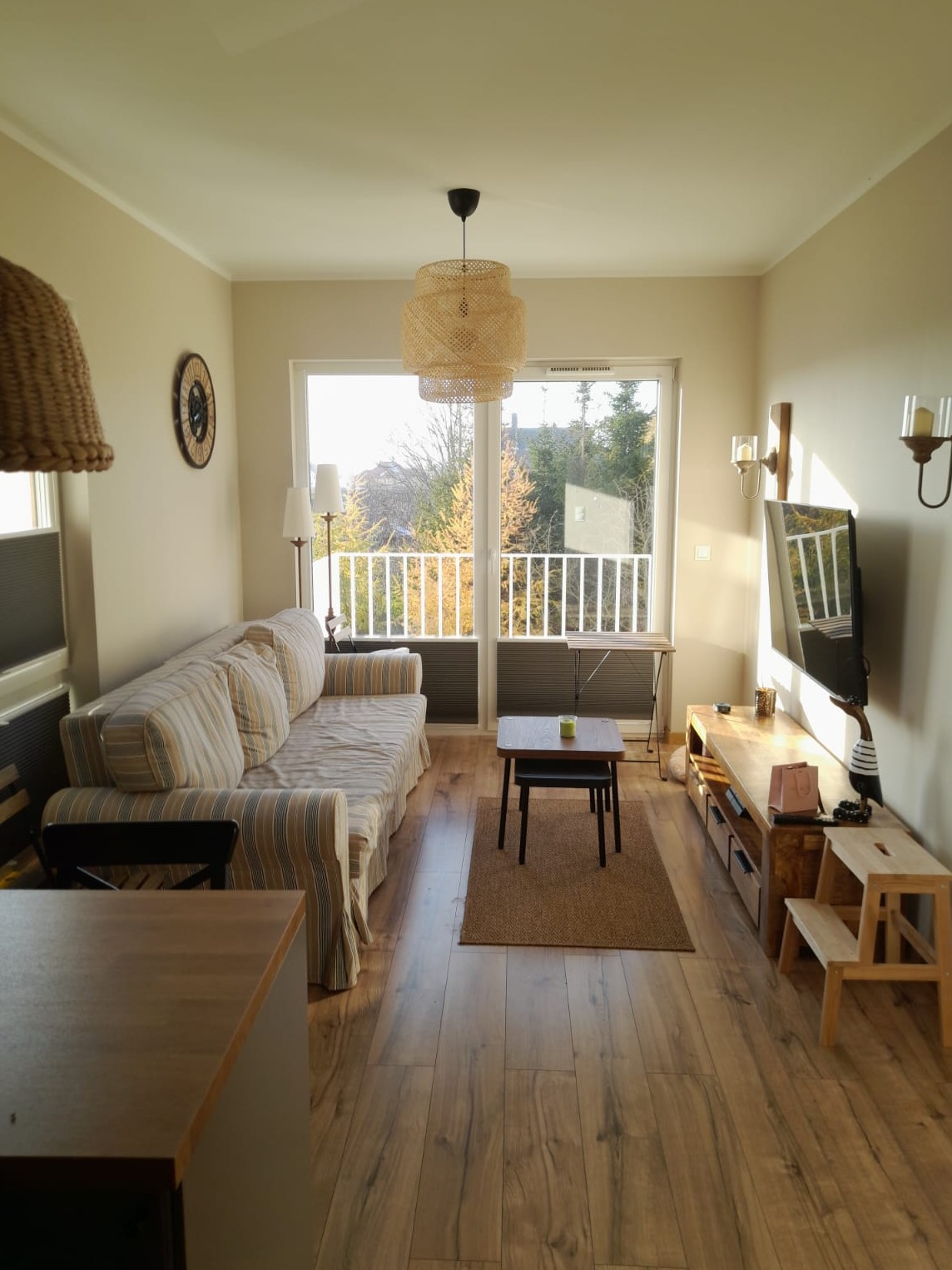 Jasne wnętrze salonu z drewnianą podłogą i balkonem. W tle widok na zieleń. Centralnie umieszczony stolik kawowy i sofa w paski. Lampa z wikliny nadaje przytulny charakter.