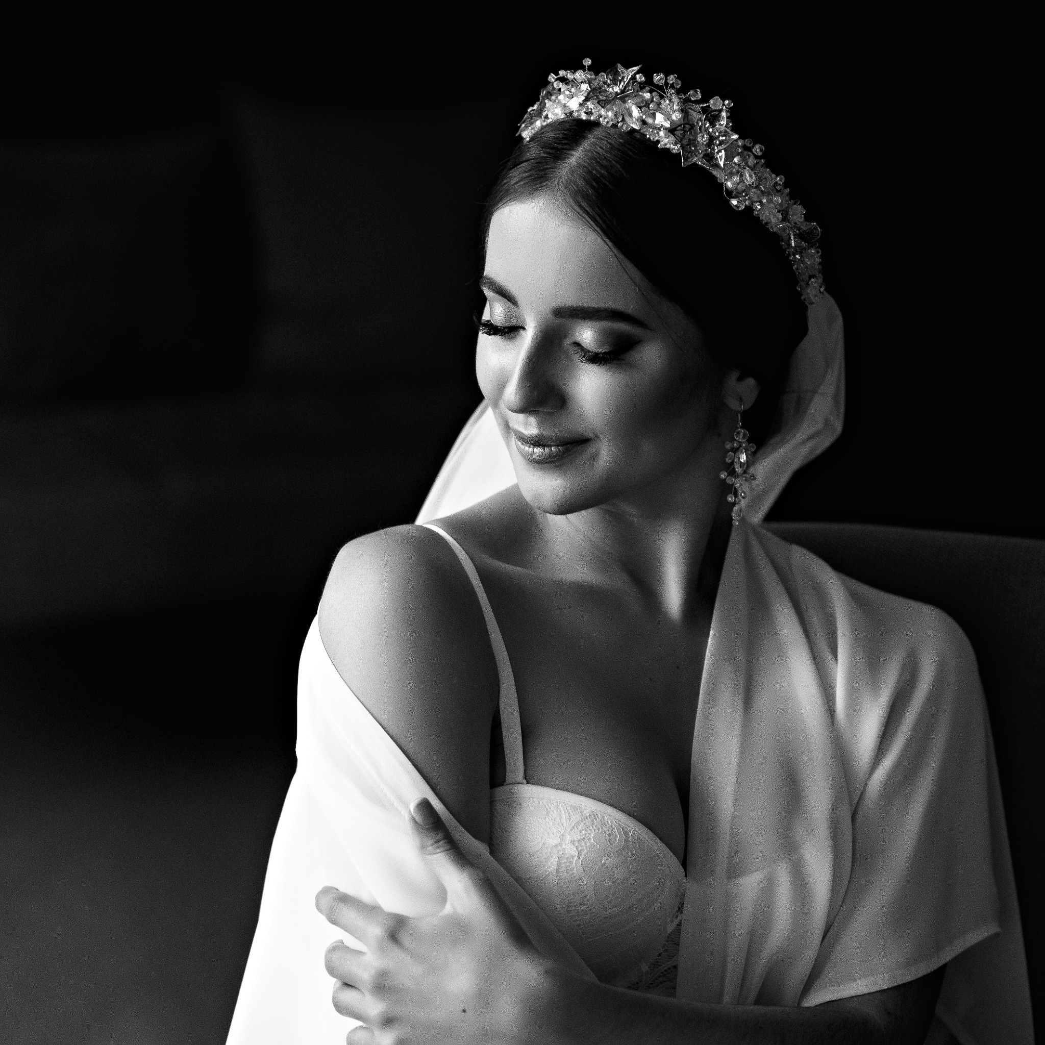 Portret w czerni i bieli panny młodej w koronie i szlafroku, z delikatnym uśmiechem i zamkniętymi oczami, przygotowującej się do ceremonii ślubnej.
