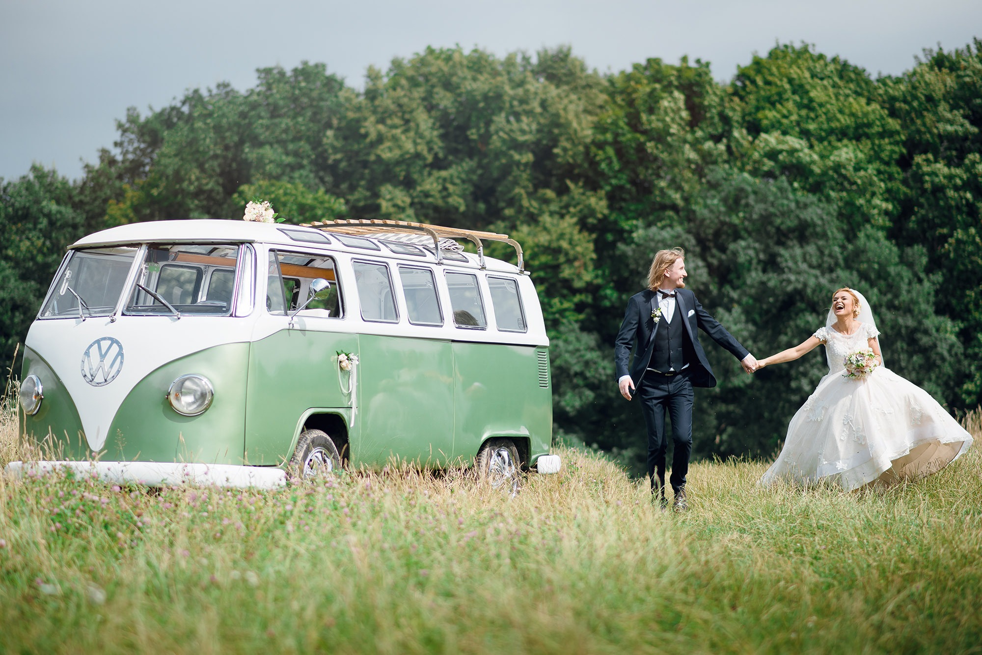 Para młoda trzymająca się za ręce, idąca po łące w kierunku zielonego, odrestaurowanego Volkswagena Transportera z dekoracjami ślubnymi, las w tle.
