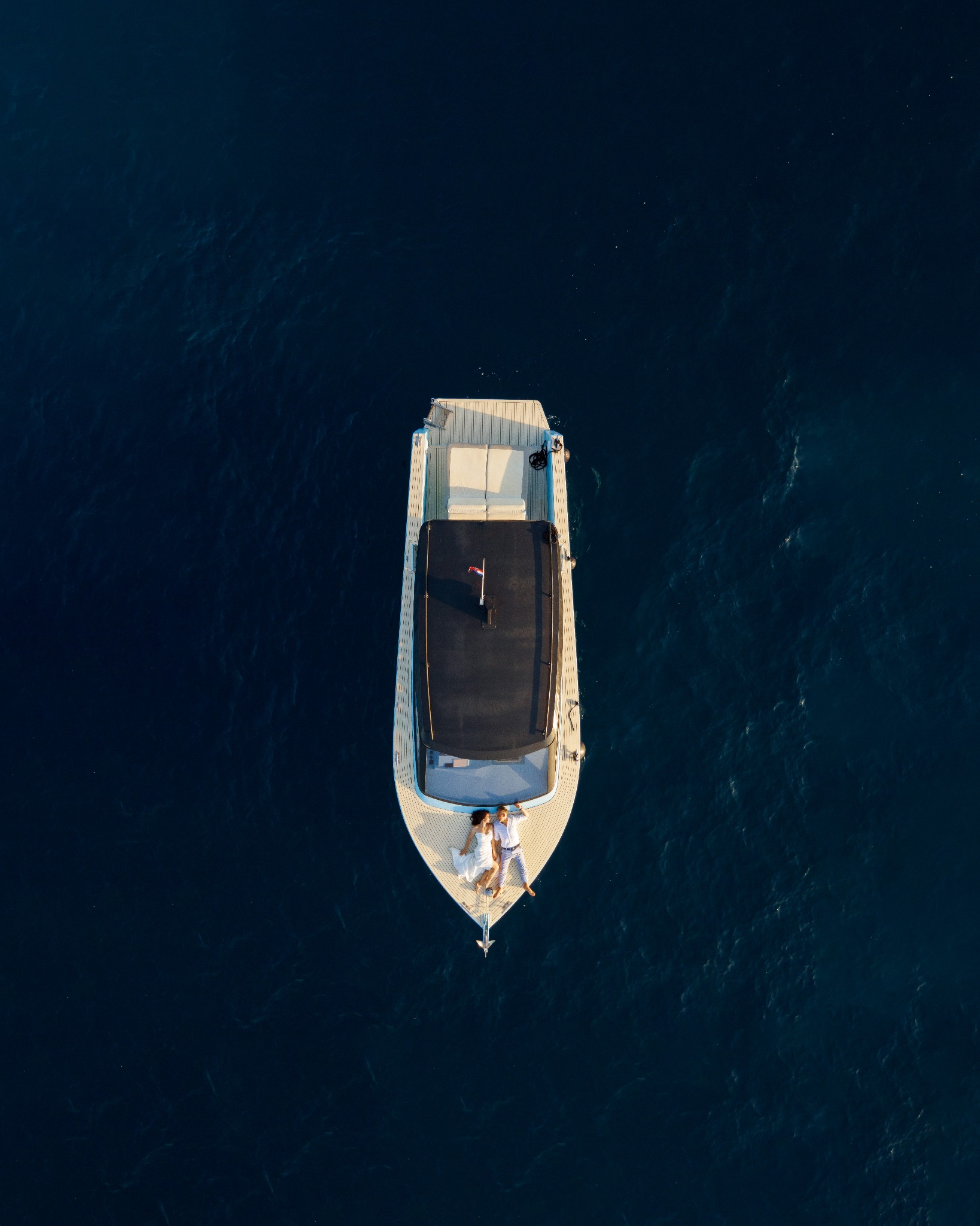 Widok z góry na parę leżącą na dziobie jachtu na tle ciemnoniebieskiej wody oceanu, kobieta w białej sukni, mężczyzna w jasnych spodniach i koszuli.