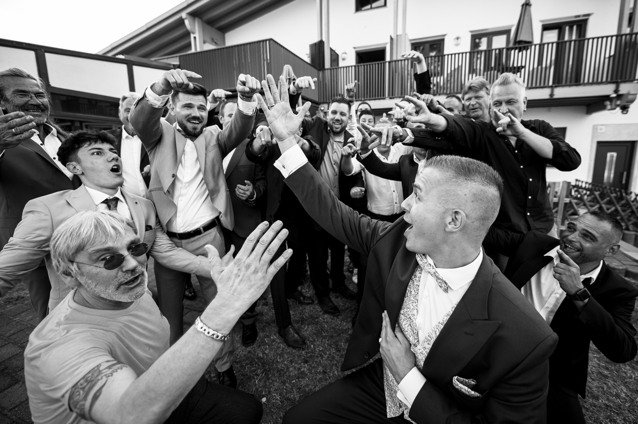 Czarno-białe zdjęcie grupy mężczyzn w garniturach, świętujących z uniesionymi rękami i wskazującymi palcami, w tle budynek z balkonem.