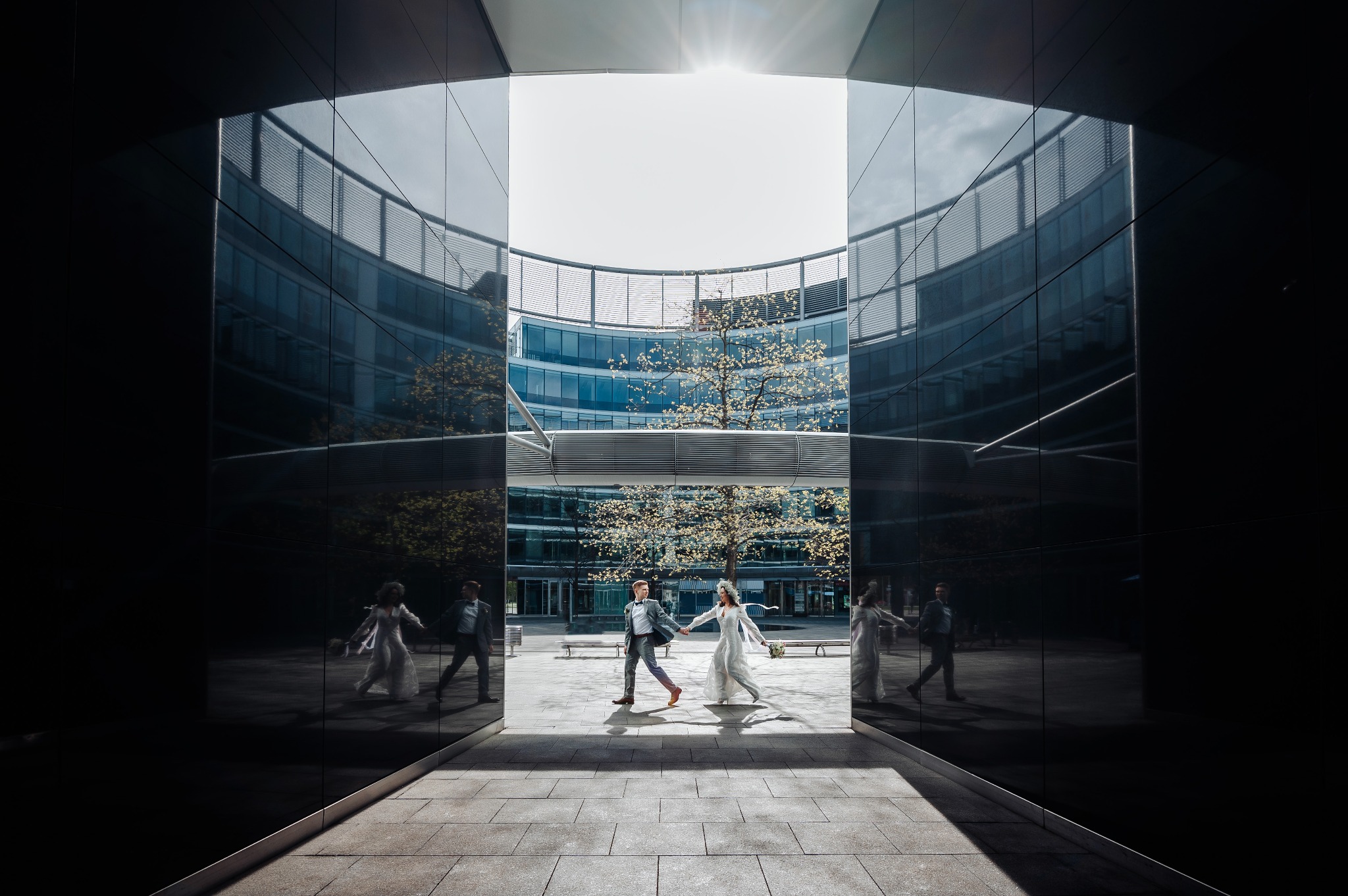 Para młoda, trzymając się za ręce, przechodzi przez nowoczesny, geometryczny korytarz z lustrzanymi ścianami, odbijającymi ich sylwetki, w tle budynek biurowy i drzewo.