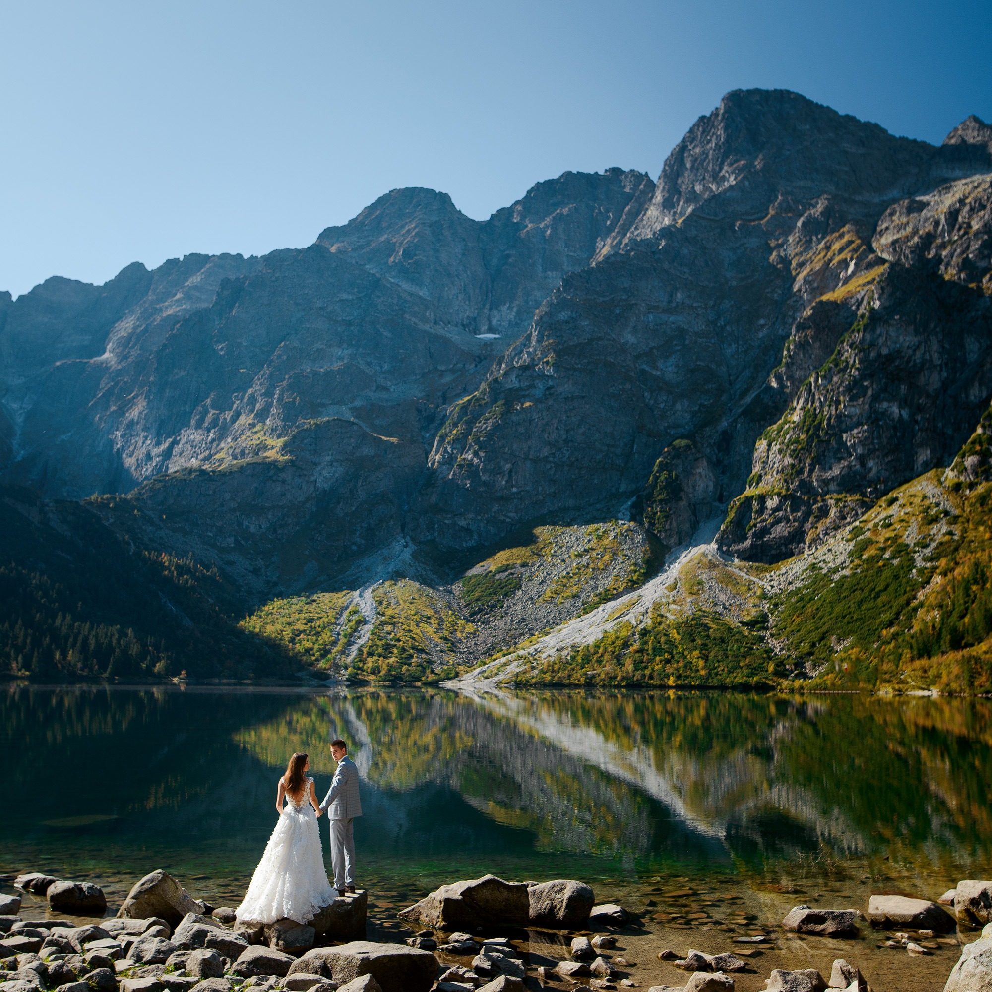 Para młoda stojąca na kamieniach nad brzegiem jeziora górskiego z odbijającymi się w wodzie szczytami, on w szarym garniturze, ona w białej sukni ślubnej z koronkowymi detalami na plecach.