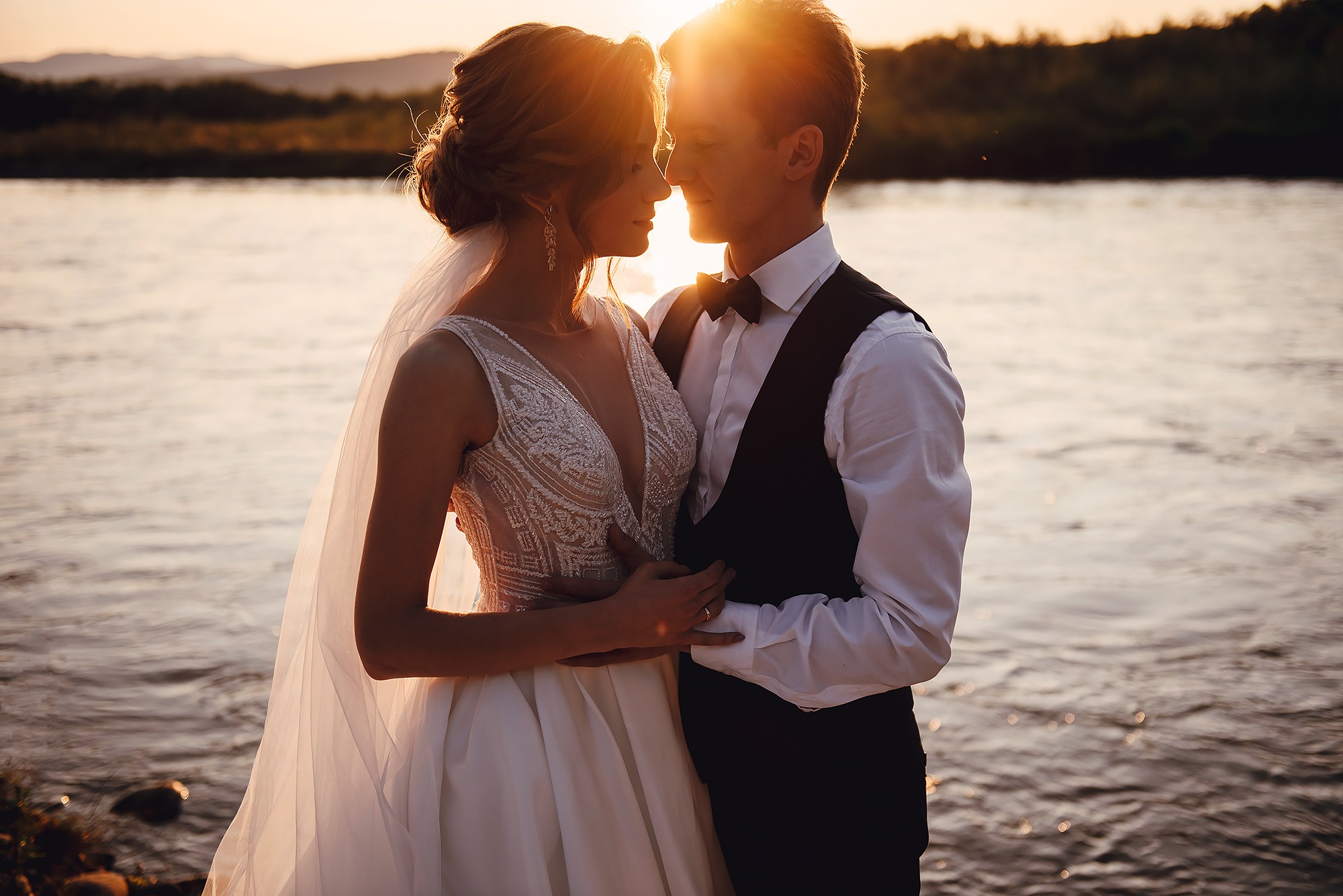 Para młoda, ona w białej sukni z koronkową górą i długim welonem, on w czarnej kamizelce i muszce, stoją blisko siebie na tle rzeki i zachodzącego słońca, z promieniami słońca padającymi na ich...