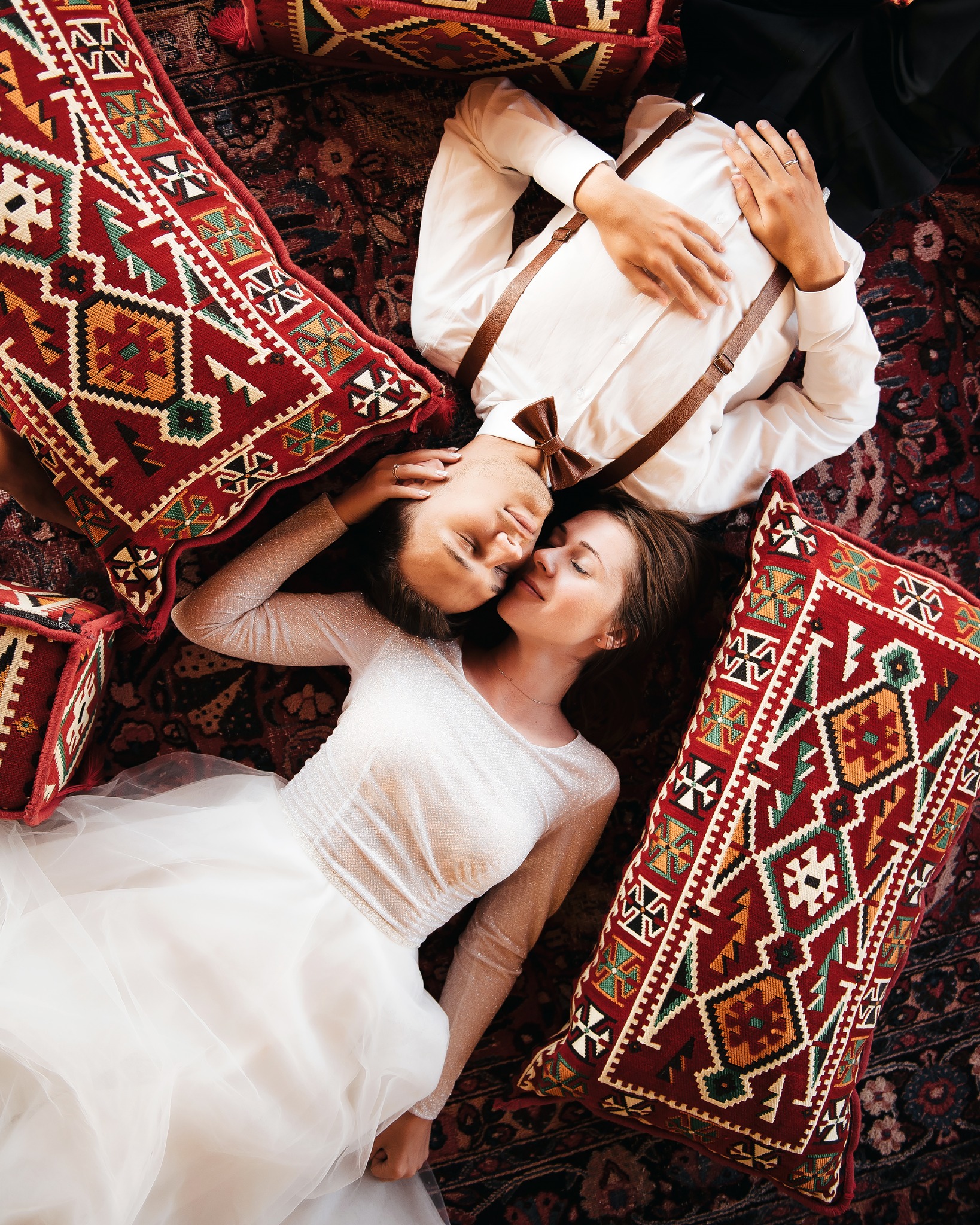 Młoda para w strojach ślubnych leży na orientalnym dywanie otoczona wzorzystymi, czerwonymi poduszkami w geometryczne wzory, panna młoda w sukni z długim rękawem, pan młody w białej koszuli...