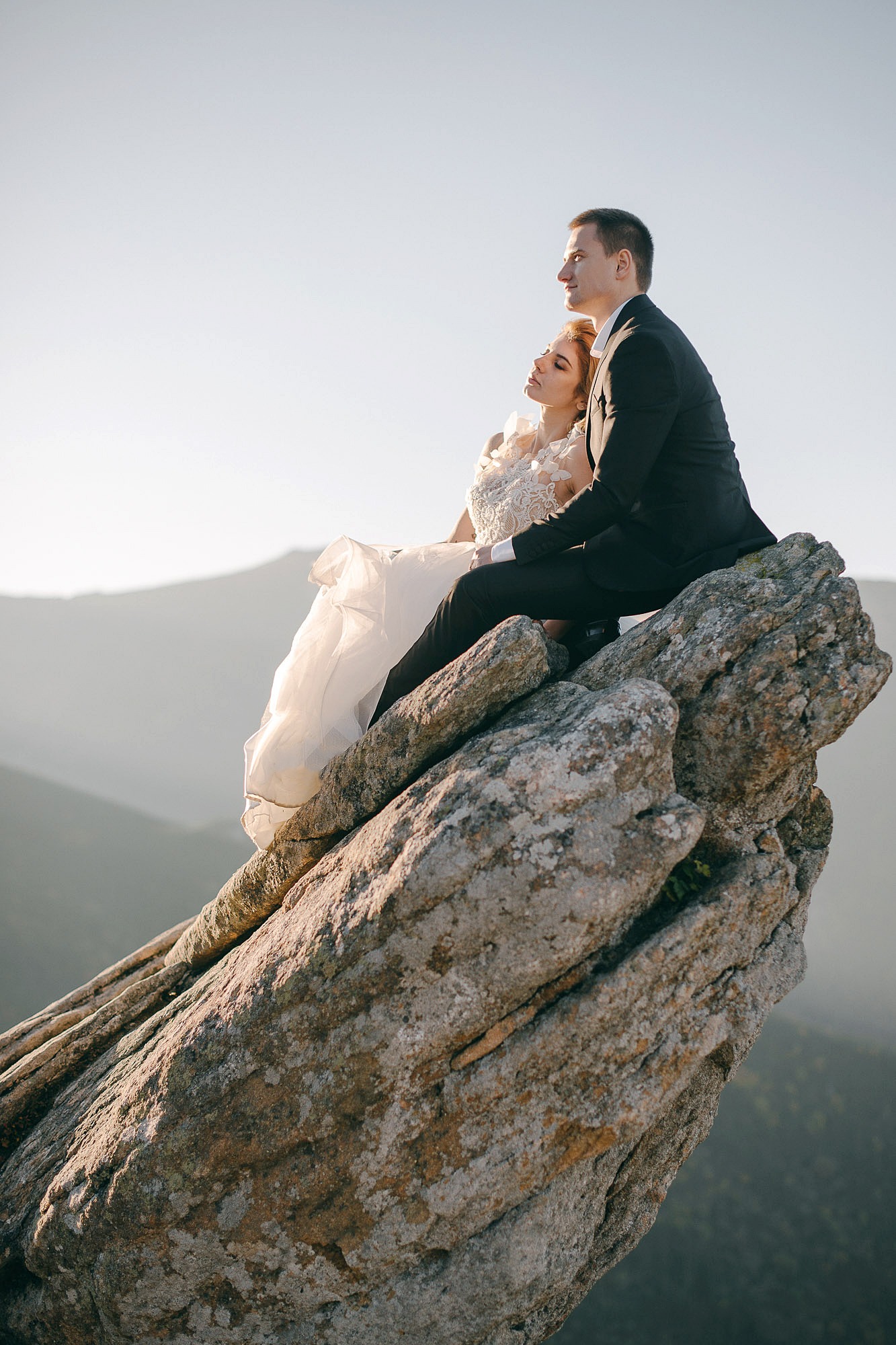 Para młoda w Krakowie siedzi na skalnym szczycie z widokiem na góry; panna młoda w białej sukni z koronkowymi aplikacjami, pan młody w czarnym garniturze, delikatne oświetlenie podkreśla naturalne...