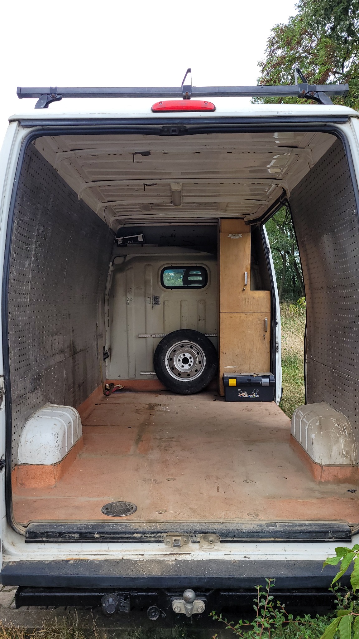 Wnętrze białego busa z zamontowaną szafką, kołem zapasowym i skrzynką narzędziową, widok z tyłu.