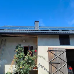 Dom z panelami słonecznymi na dachu, drabina oparta o ścianę, drewniane wrota stodoły otwarte, widoczny samochód w środku, krzew róży pnącej przy oknie.