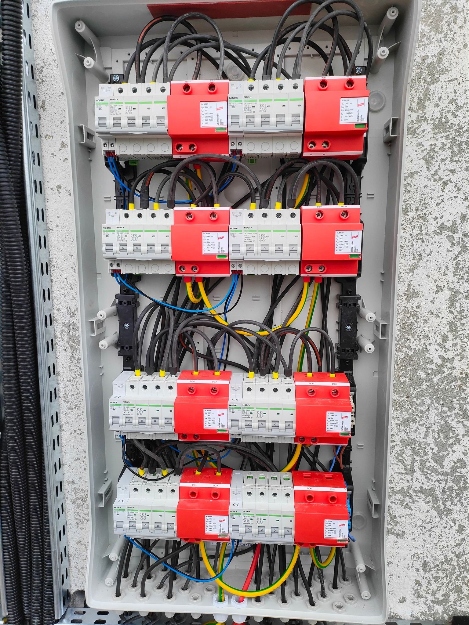 Rozdzielnica elektryczna z zabezpieczeniami przepięciowymi DC, widoczne przewody w kolorach żółto-zielonym, niebieskim i czarnym, zamontowana na szarej ścianie.