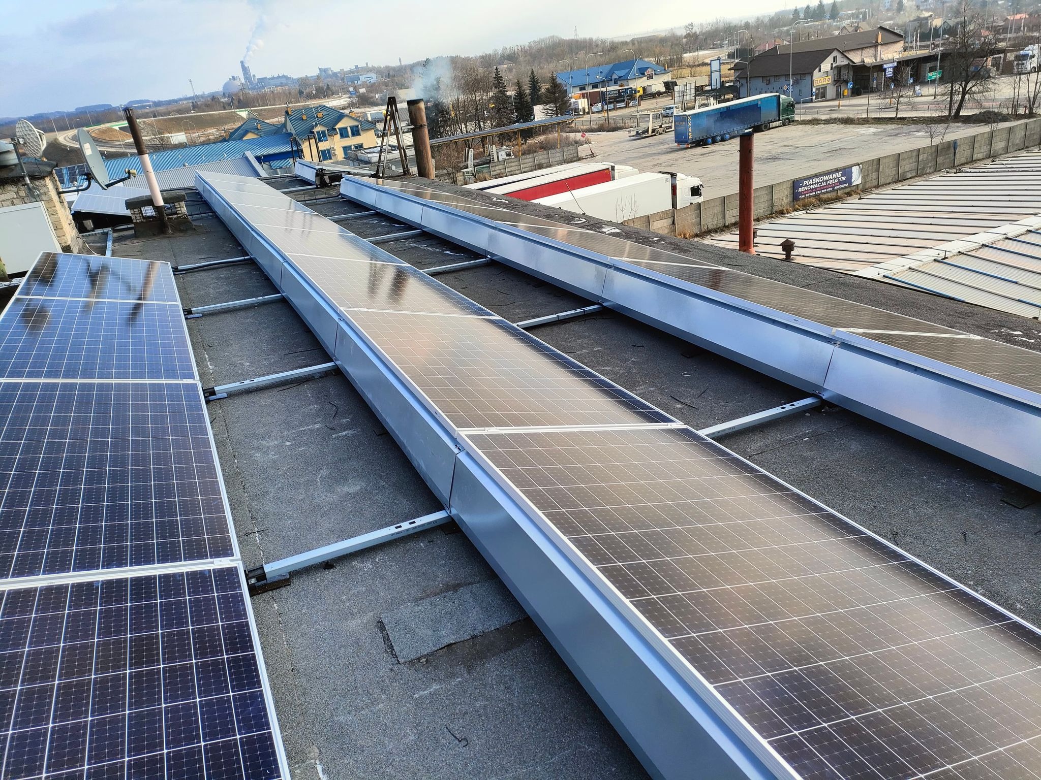 Rząd paneli słonecznych z aluminiową ramą na płaskim, szarym dachu budynku przemysłowego, w tle komin fabryczny i zabudowa miejska.